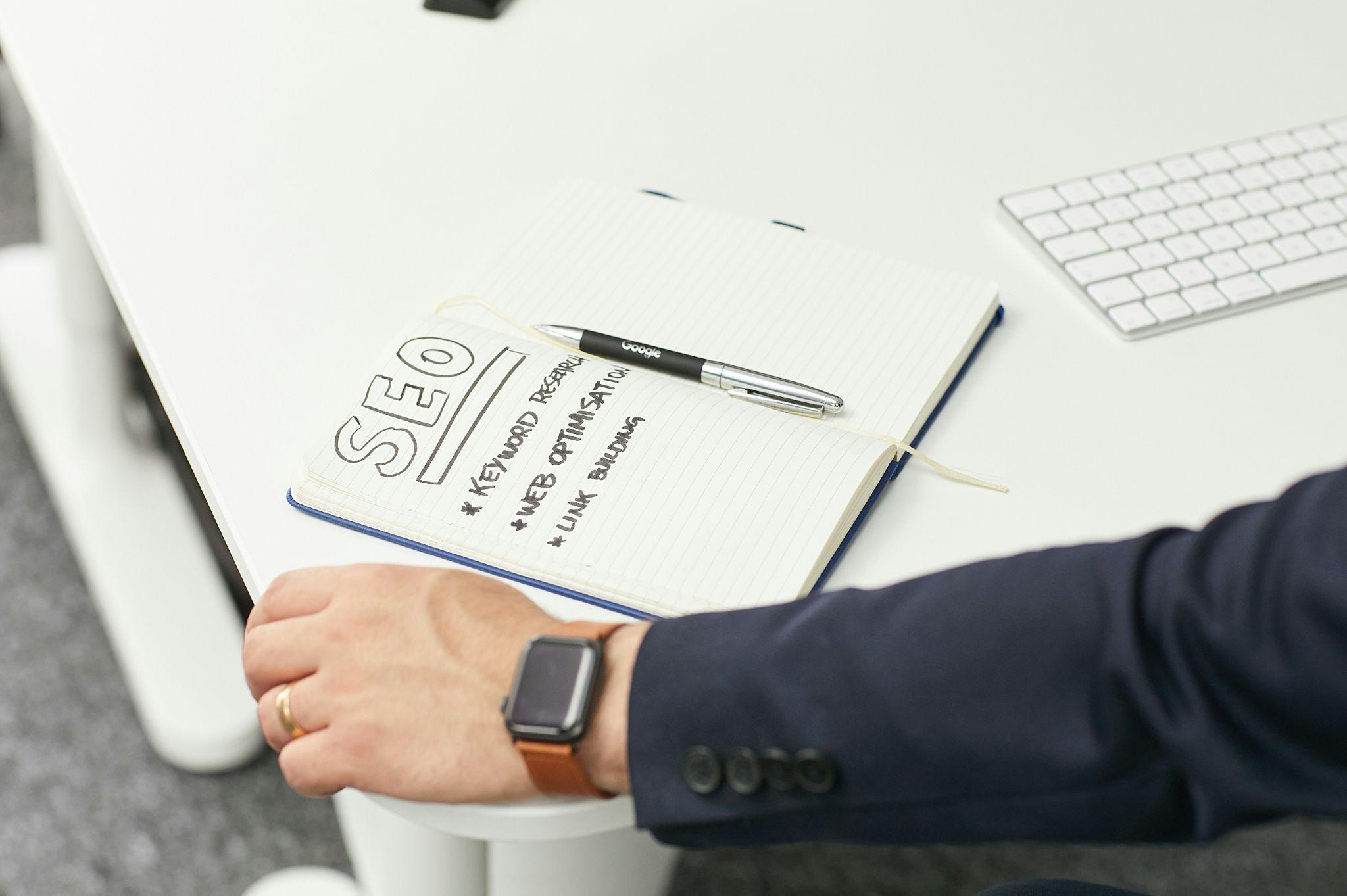 Notebook open to an SEO planning page with keyword ideas, a pen resting on the page, and a person wearing a smartwatch nearby on a white desk.