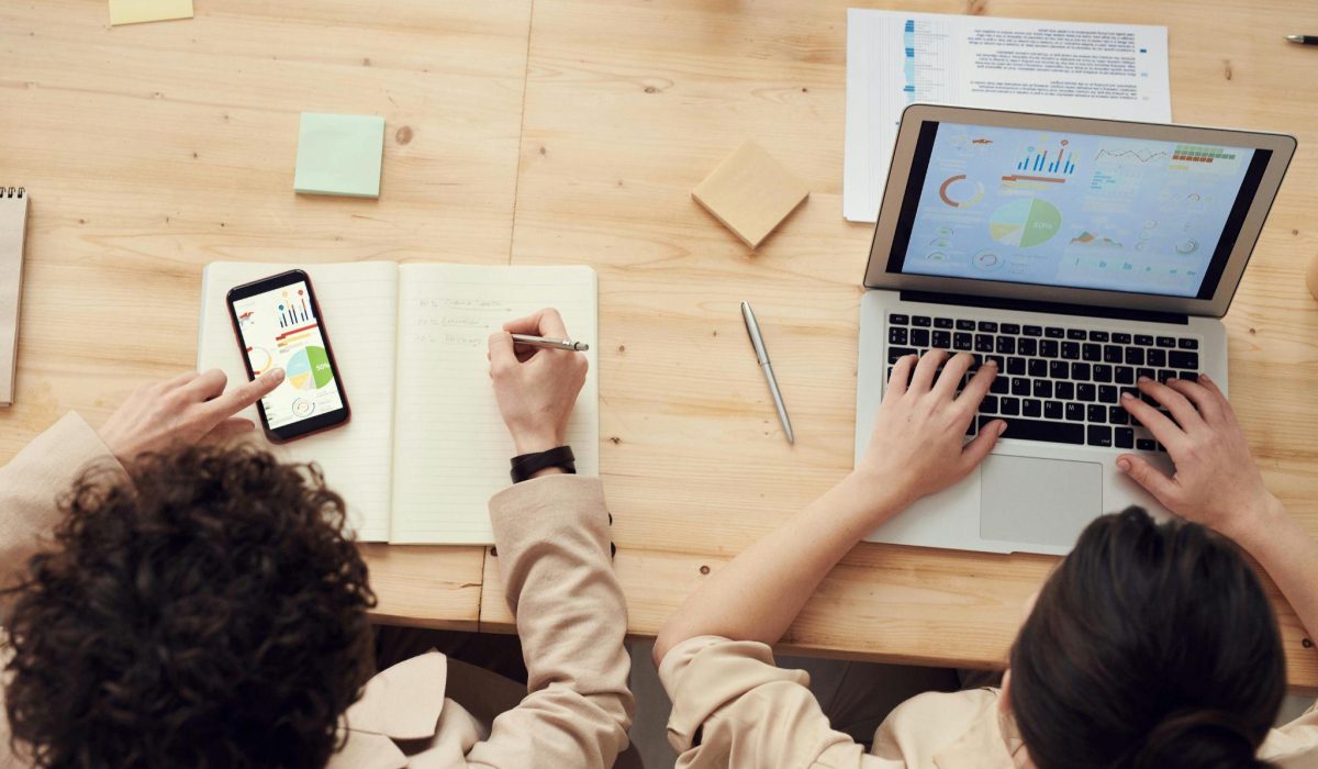 Two people work at a light wooden table: one types on a laptop with charts on screen while the other writes in a notebook beside a smartphone displaying graphs and sticky notes around.
